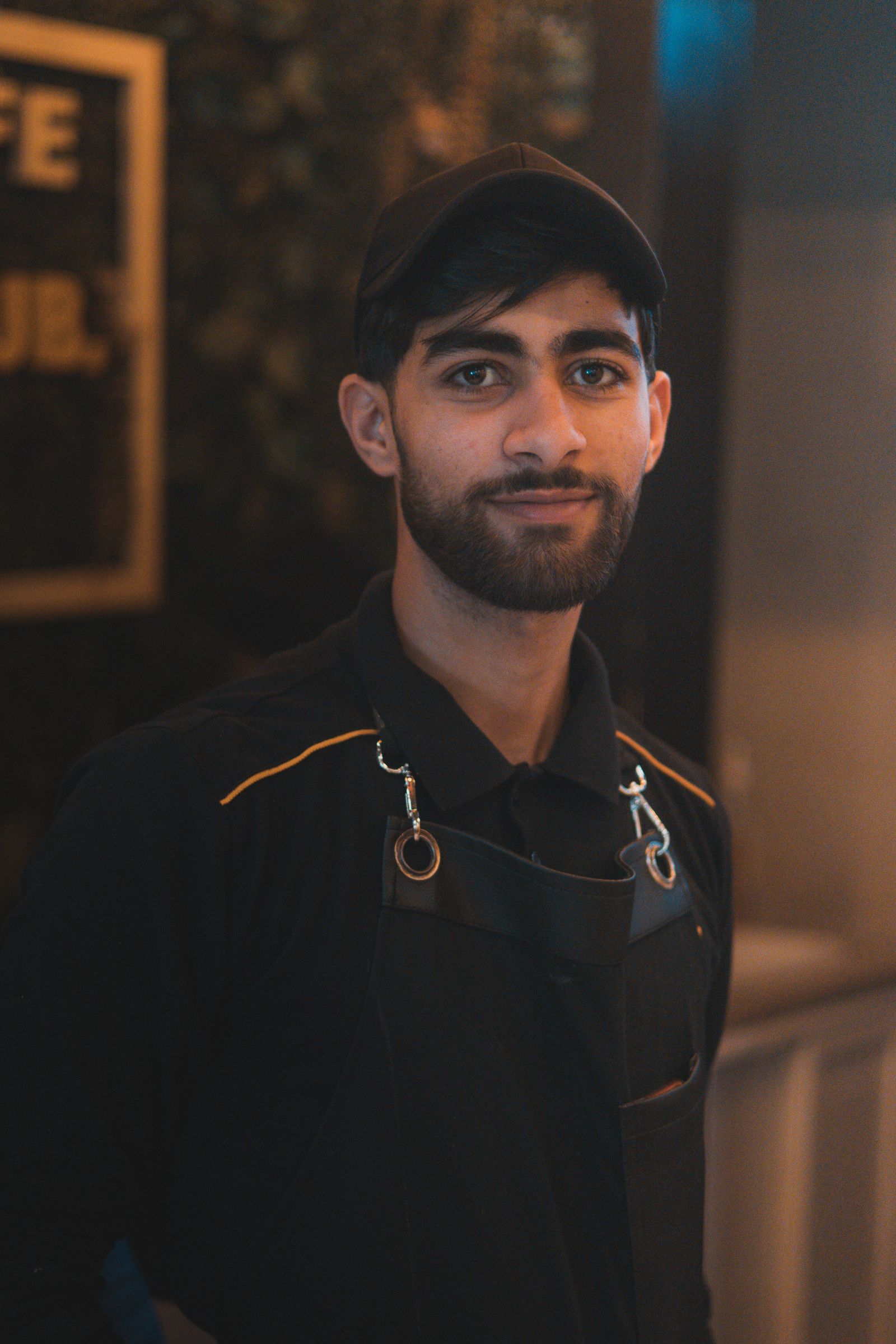 Portrait of a young male barista wearing a cap and apron in a coffee shop setting.