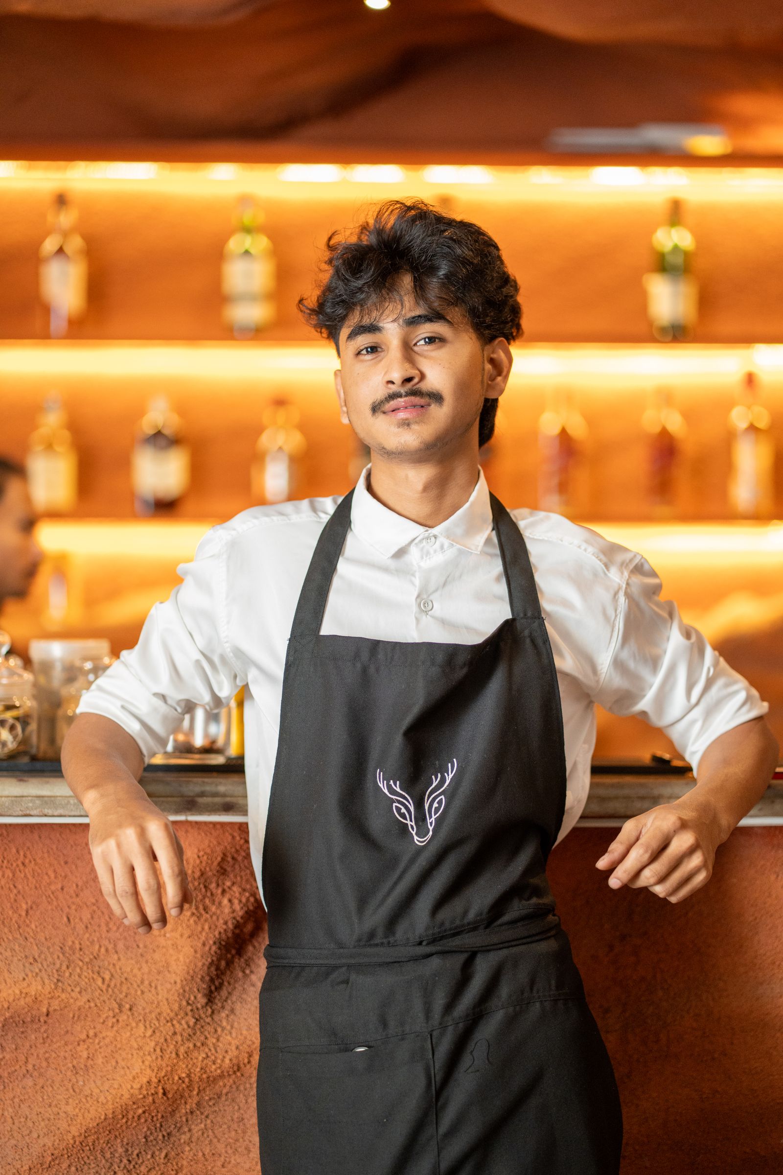 Young bartender posing confidently against a sleek bar backdrop with illuminated shelves.